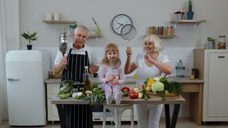 Senior-woman-and-man-with-grandchild-girl-making-a-funny-dance-with-strainer-and-vegetables-at-home