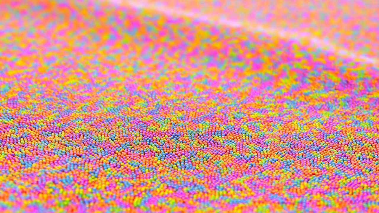Dense layers of small colorful spheres rise and fall into gentle ridges as the patterned surface shifts over time, with vivid warm and cool tones fading softly toward the background.