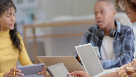 Diverse-group-of-teenage-friends-studying-at-table-with-tablets-and-laptops-at-home,-slow-motion