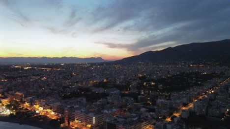 aerial push, tilt down high angle view of night cityscape of kalamata, peloponnesus, greece, beautiful sunset in the background on a cloudy afternoon 4k