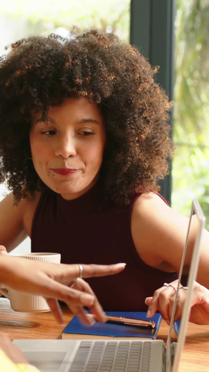 Vertical video: Sipping woman in maroon top by window reviewing laptop while companion pointing