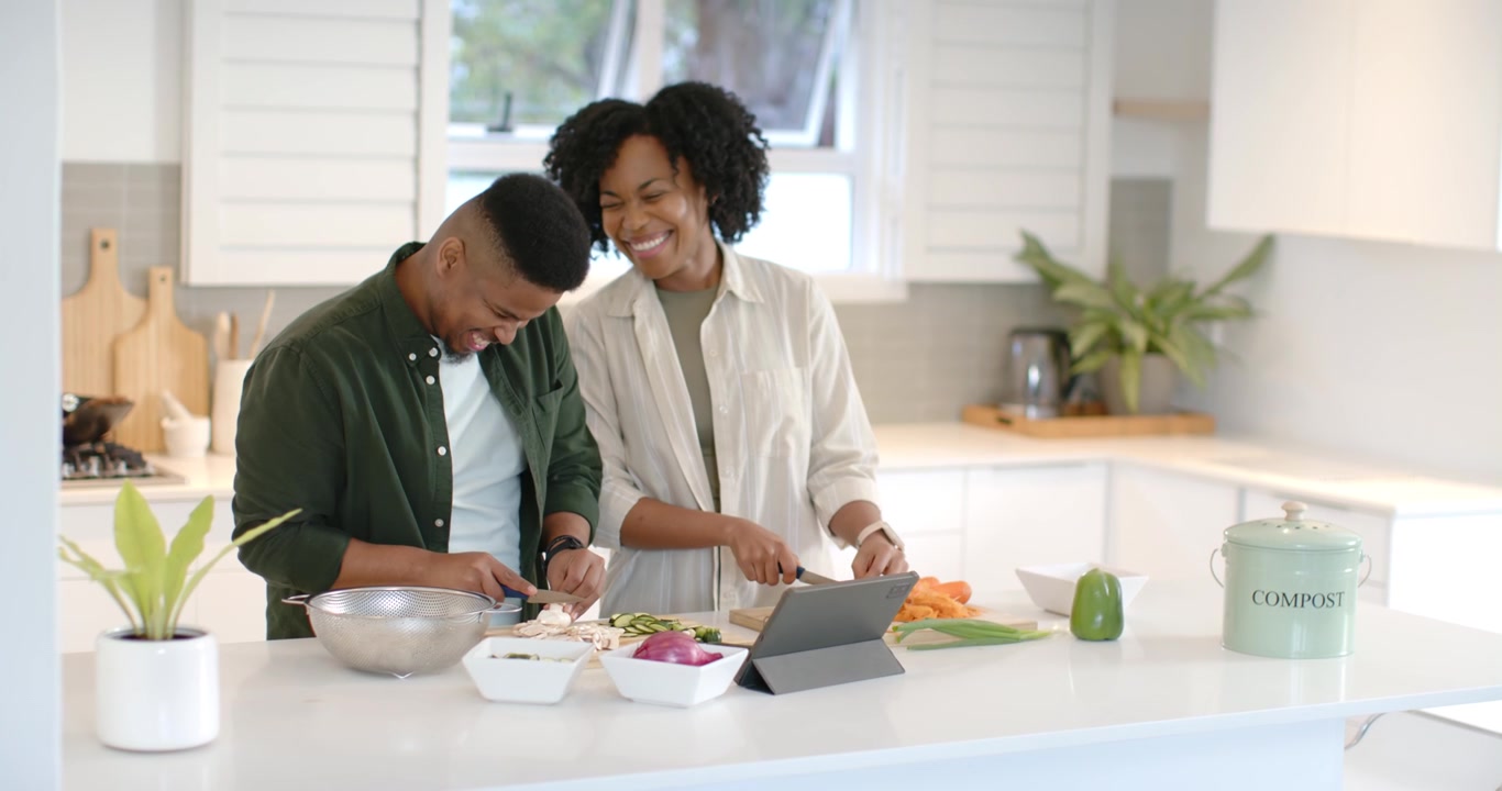 Una coppia afroamericana segue una ricetta sul tablet, taglia ingredienti sui taglieri e si scambia sguardi.