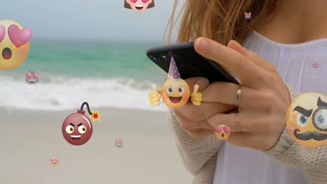 animation of emoticons over hands of caucasian woman using smartphone at beach