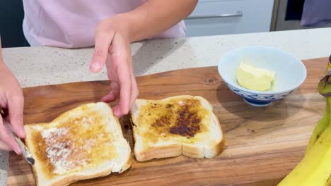 person prepares toast by spreading butter and dark yeast extract on sliced bread in a brightly lit modern kitchen, using a butter knife and wooden board