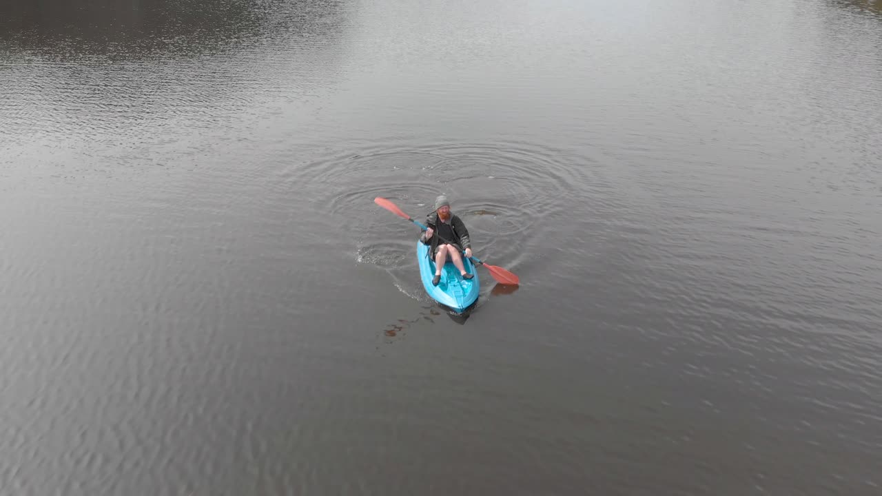 toma aérea rastreando a un hombre barbudo desde el frente mientras está en un kayak azul sentado en la parte superior de un lago