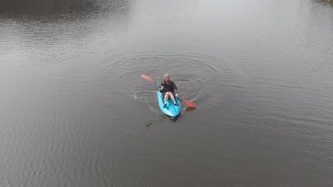 aerial shot tracking a bearded man from the front while he is on a blue sit on top kayak on a lake