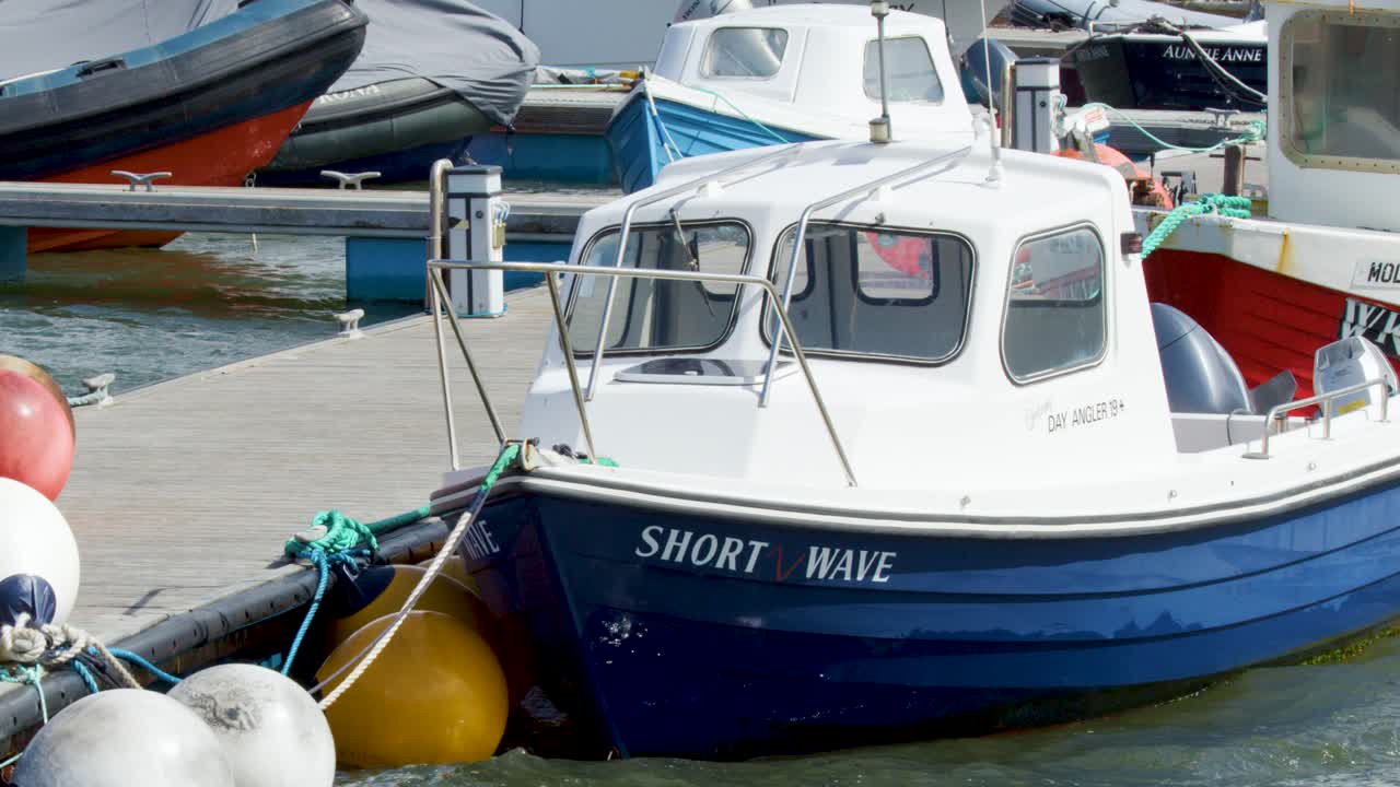 A blue and white fishing boat moves slowly toward a dock lined with colorful buoys and moored vessels under bright daylight, camera tracking forward