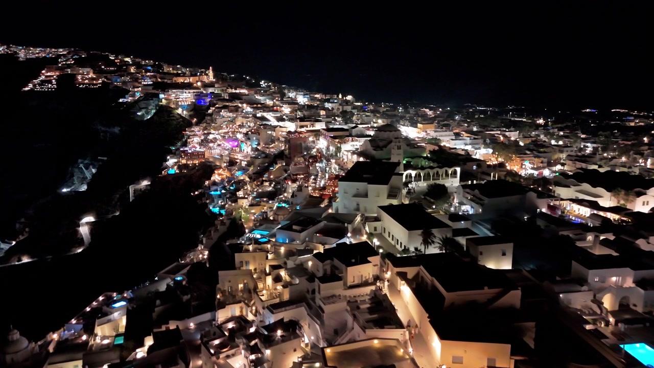 Le affollate strade collinari di Fira risplendono con ristoranti, bar e piscine illuminate mentre la telecamera sorvola il villaggio di notte