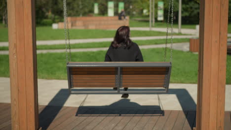 back view of woman on wooden swing chair swinging with hair fluttering in wind, surrounded by greenery in tranquil park setting, trees in background, blurred figure moving