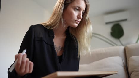 Young,-tattooed-blonde-woman-working-and-studying-effortlessly-from-her-home-sofa,-taking-notes-and-immersed-in-a-laptop,-blending-comfort-with-productivity