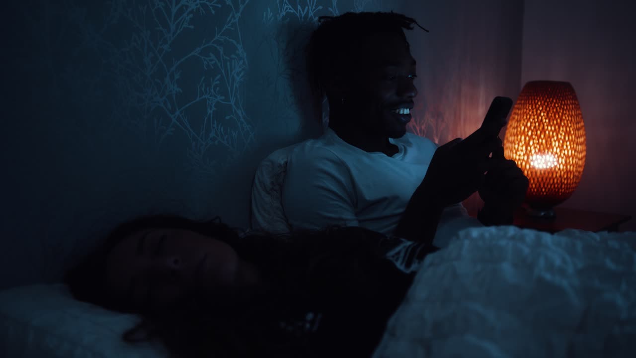 Un uomo sorride e si volta verso il partner mentre sono a letto di notte, usando uno smartphone.