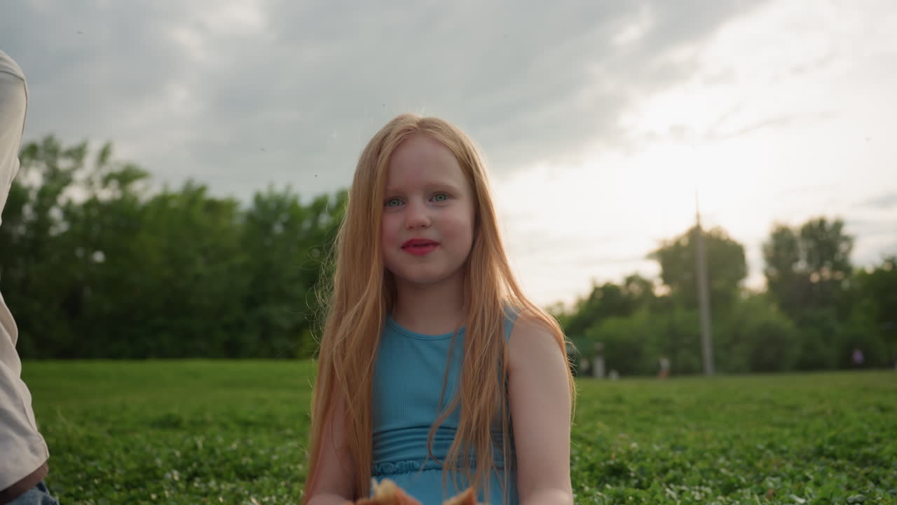 portrait teenage girl waving pastry wrapped in brown paper in sunny recreational park smiling at camera while enjoying outdoor snack time with blurred trees background summer leisure