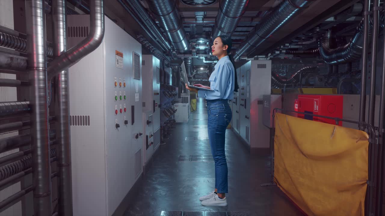 Full Body Side View Of An Asian Female Worker Standing In Engine Control Room, Observes By Looking Up Before She Come To Concentrating On The Laptop And Keep On Typing