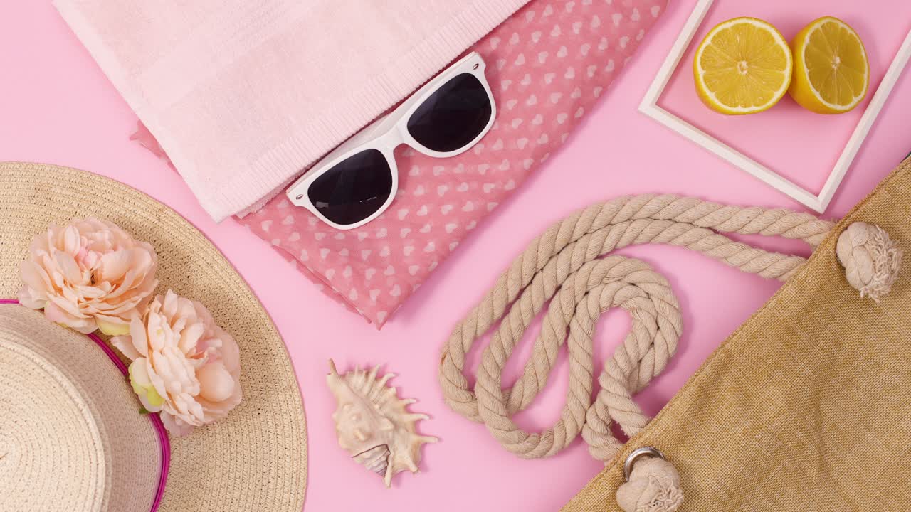 sombrero de paja de verano con flores y accesorios de playa aparecen sobre un fondo rosa pastel.