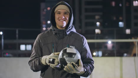 atleta sonriente con capucha y guantes sosteniendo una pelota de fútbol en un campo deportivo urbano por la noche, edificios de gran altura iluminados, postes de portería y luces bokeh vibrantes