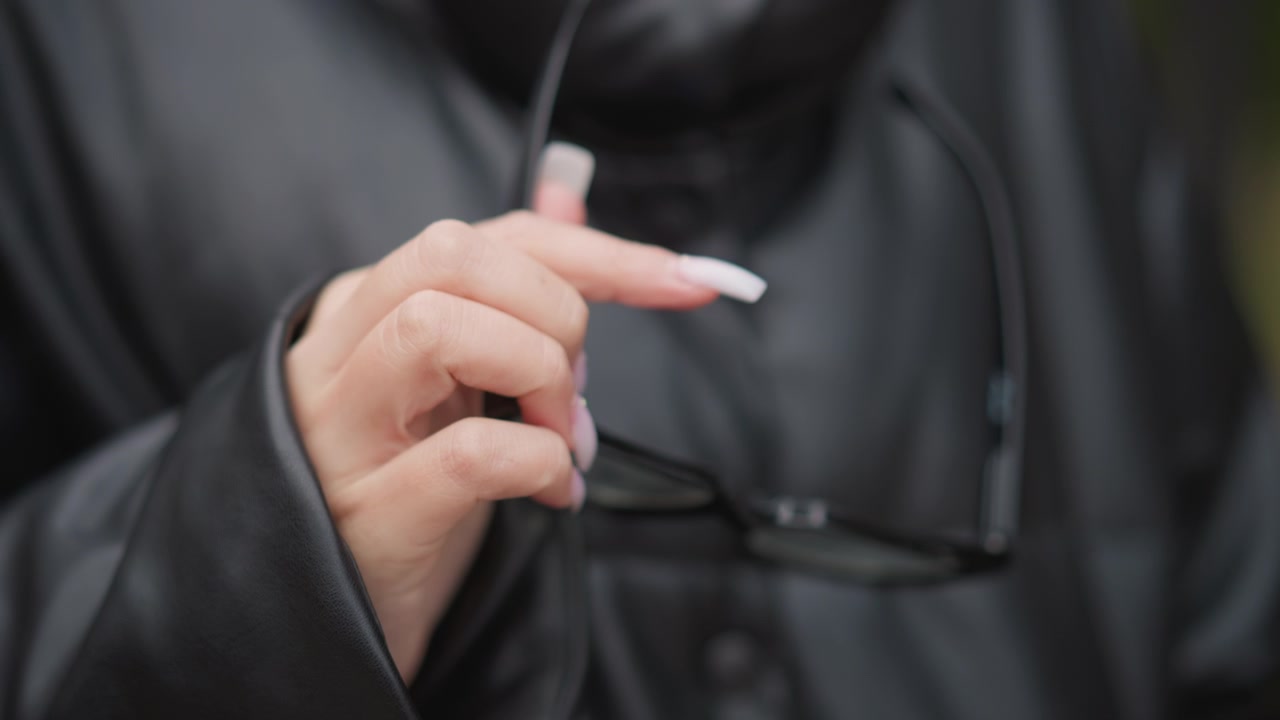 Primo piano della mano di una donna con unghie lunghe che tiene occhiali neri su uno sfondo sfocato. La mano rimane ferma per tutto il tempo, enfatizzando la lunghezza e la texture delle unghie mentre afferra gli occhiali.
