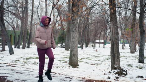 young woman in winter outfit warms up in cold outdoor park, walking on snowy ground, dressed in hoodie, boots, and gloves, she moves through scenic park surrounded by leafless trees