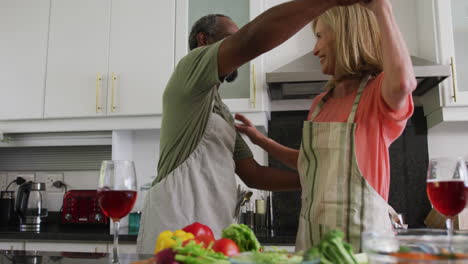 Diversa-Pareja-De-Ancianos-Usando-Delantales-Bailando-Mientras-Preparan-Comida-En-La-Cocina.