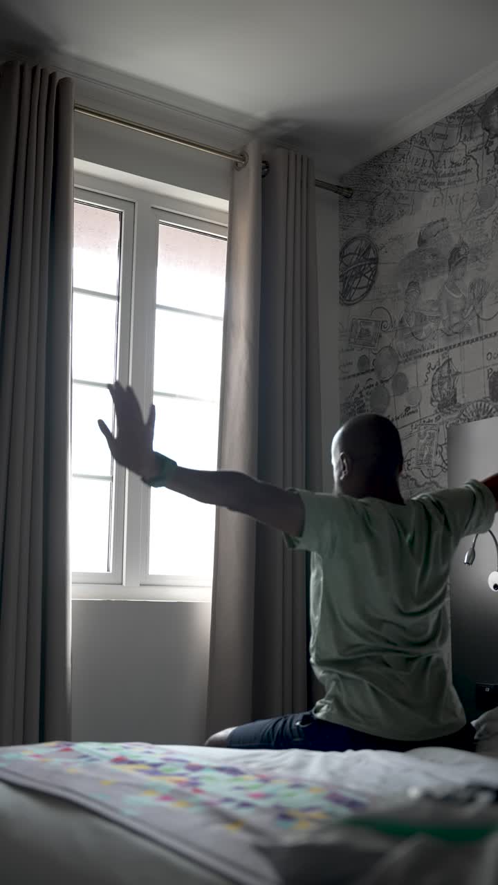 un hombre se sienta en una cama en una habitación de hotel, estirándose hacia la ventana brillante, transmitiendo relajación y estado de ánimo de viaje. video vertical