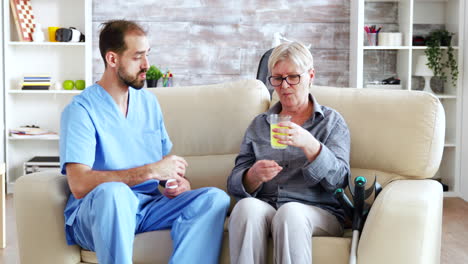 Male-doctor-sitting-on-sofa-with-senior-woman-in-nursing-home-giving-her-daily-pills.