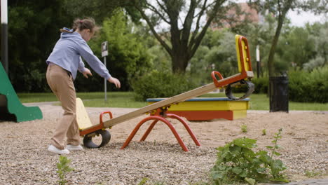 Seitenansicht-Eines-Kleinen-Mädchens-Mit-Down-Syndrom-Und-Ihrer-Blonden-Freundin,-Die-An-Einem-Windigen-Tag-Auf-Einer-Holzwippe-Im-Park-Reitet.-Dann-Beginnen-Sie-Zu-Schwingen