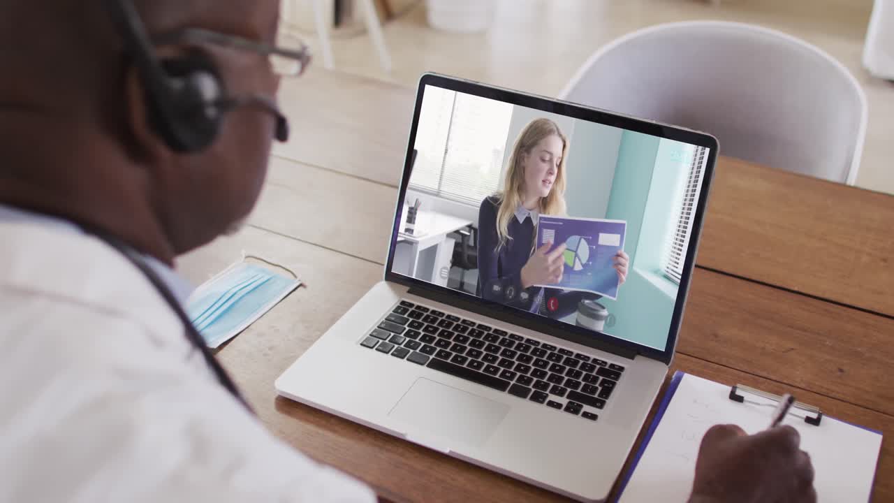 Afroamerikansk mandlig læge med headset på, der tager noter under et videoopkald på sin bærbare computer