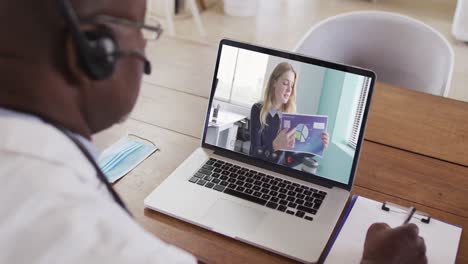 African-american-male-doctor-wearing-phone-headset-taking-notes-while-having-a-video-call-on-laptop