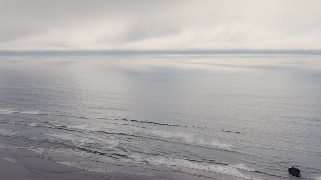 Cinematic aerial drifting above the Pacific Ocean at Arcadia Beach on the Oregon Coast as gentle waves roll in beneath low clouds with a thin blue line of sky glowing on the distant horizon