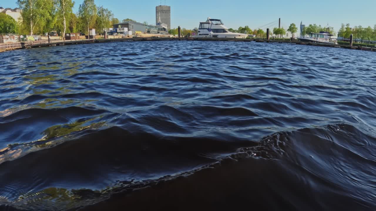Vista dal basso di increspature sull'acqua vicino ad Andrejosta a Riga, che catturano la luce del sole, dettagli del porto, yacht e silhouette industriali in una luminosa atmosfera costiera