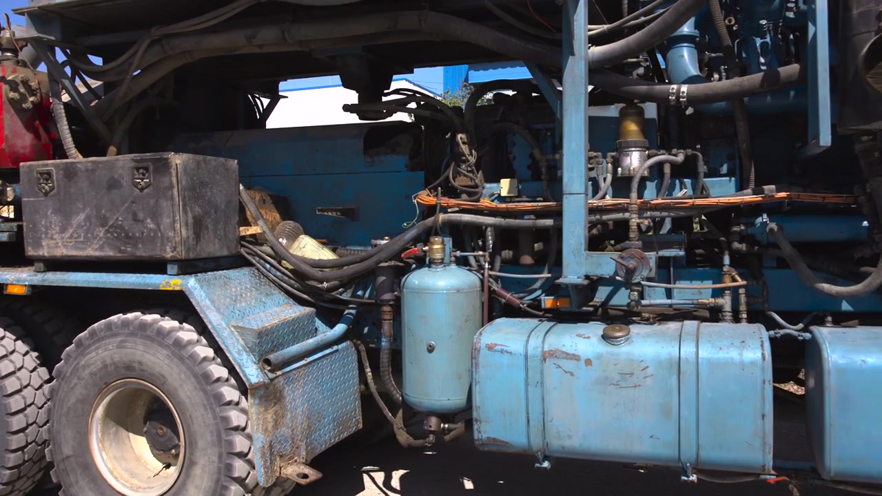 Close side view of heavy industrial truck shows exposed engine parts, thick hoses, black housings, fuel tanks, and dual rugged tires beside blue and yellow structure, with hard daylight, worn metal