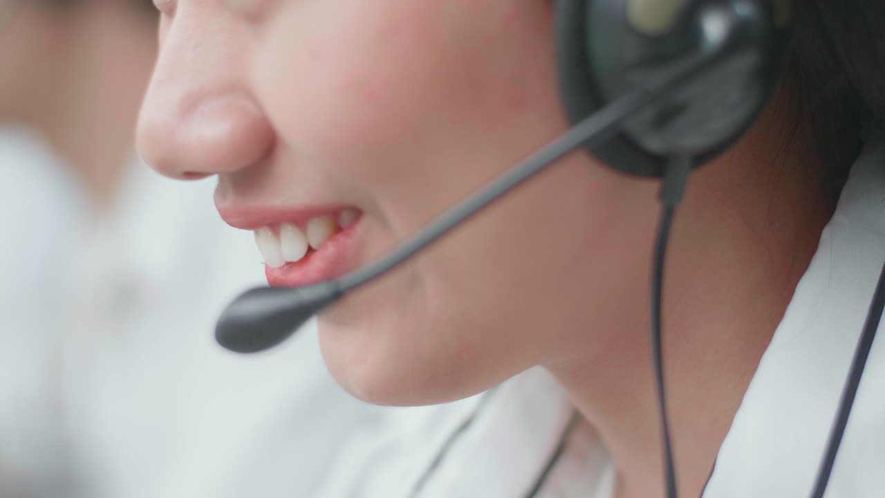 Close Up Of An Asian Woman Call Center Agent Wearing Headset Smiling While Speaking To Customer On The Call During Working With Her Colleagues At The Office