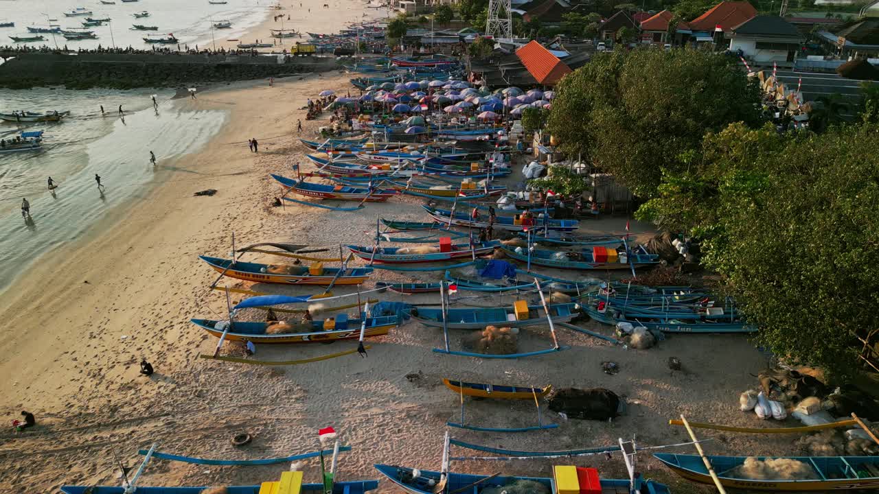 Aerial video of Jimbaran beach Bali shows traditional fishing canoes on the shore, waves touching golden sand, crowds enjoying coastal scenery, and unique tropical culture blending with seafront views