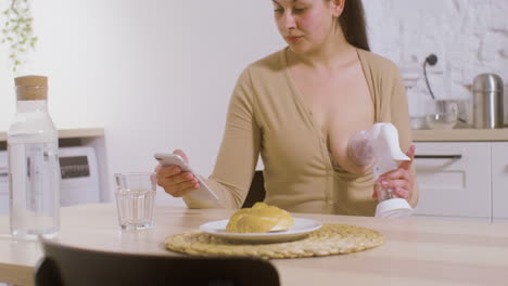 young mother using breast pump and scrolling on mobile phone while sitting at table at home