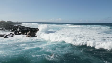 Low-flying-aerial-view-of-the-ocean-waves-crashing-on-a-seawall-on-a-sunny-day