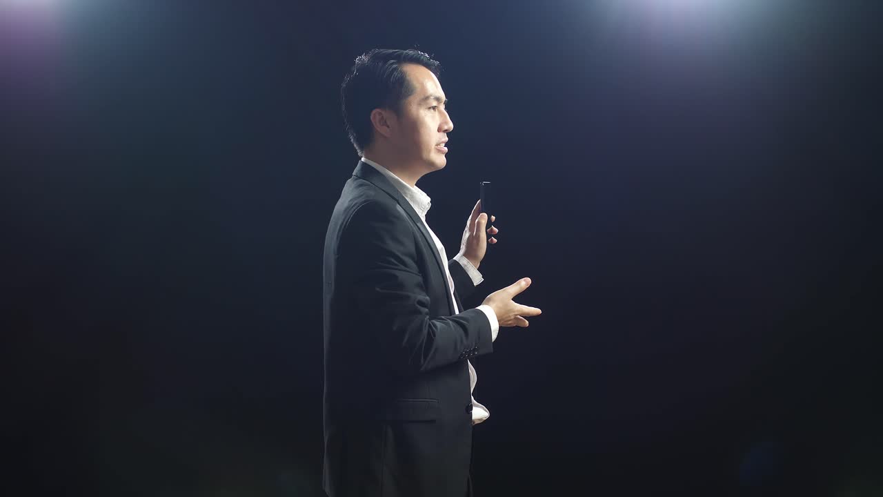 블랙 스크린 스튜디오에서 말하는 동안 스마트폰을 들고 가리키는 비즈니스 슈트 남자
