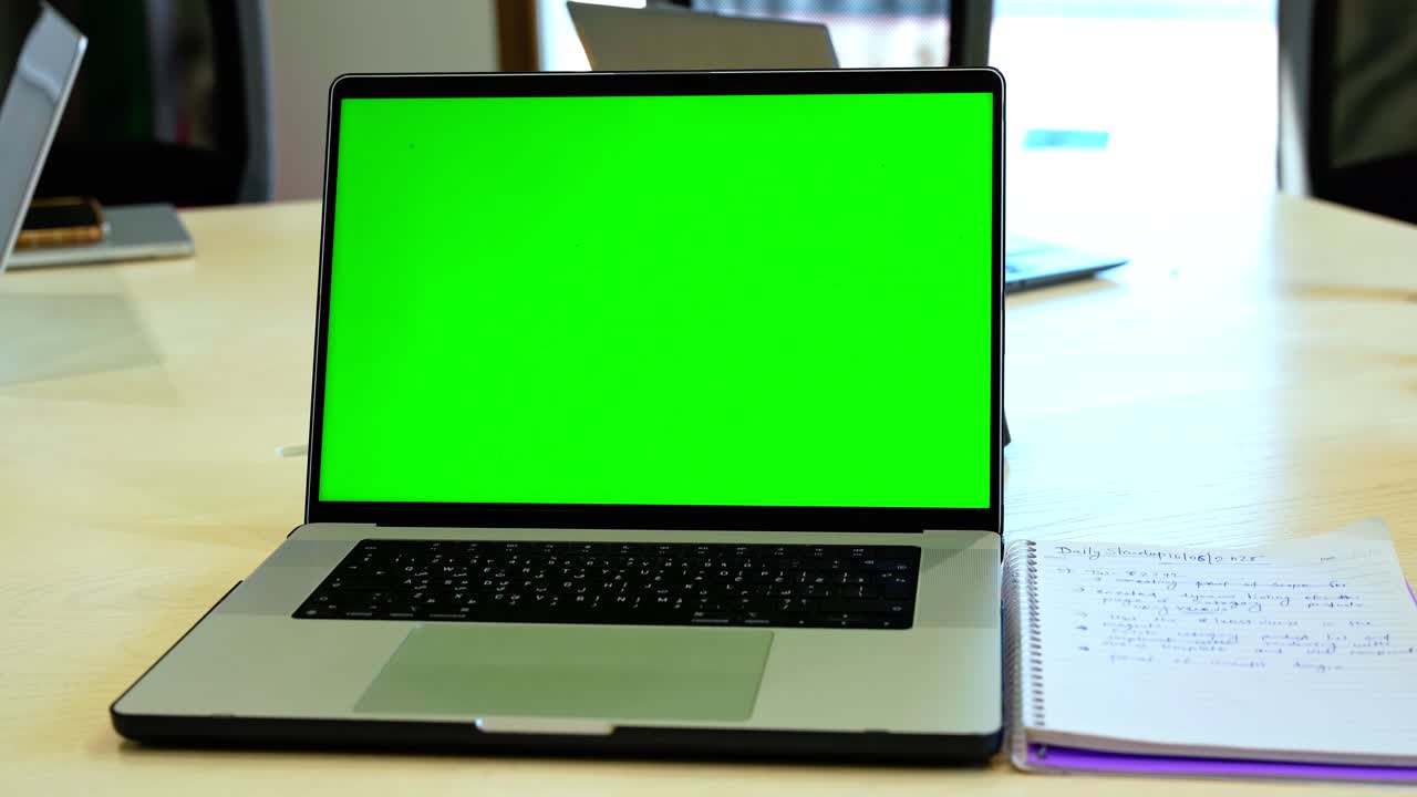 Laptop with green screen sits on a desk beside notebook and tech devices in a bright office. Ideal for template insertions, business B-roll, or modern workspace scenes.
