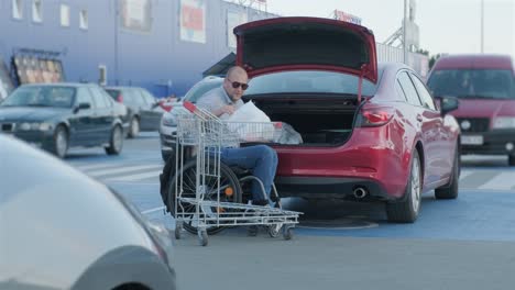 adult man with disabilities in a wheelchair puts purchases in the trunk of a car in a supermarket parking lot