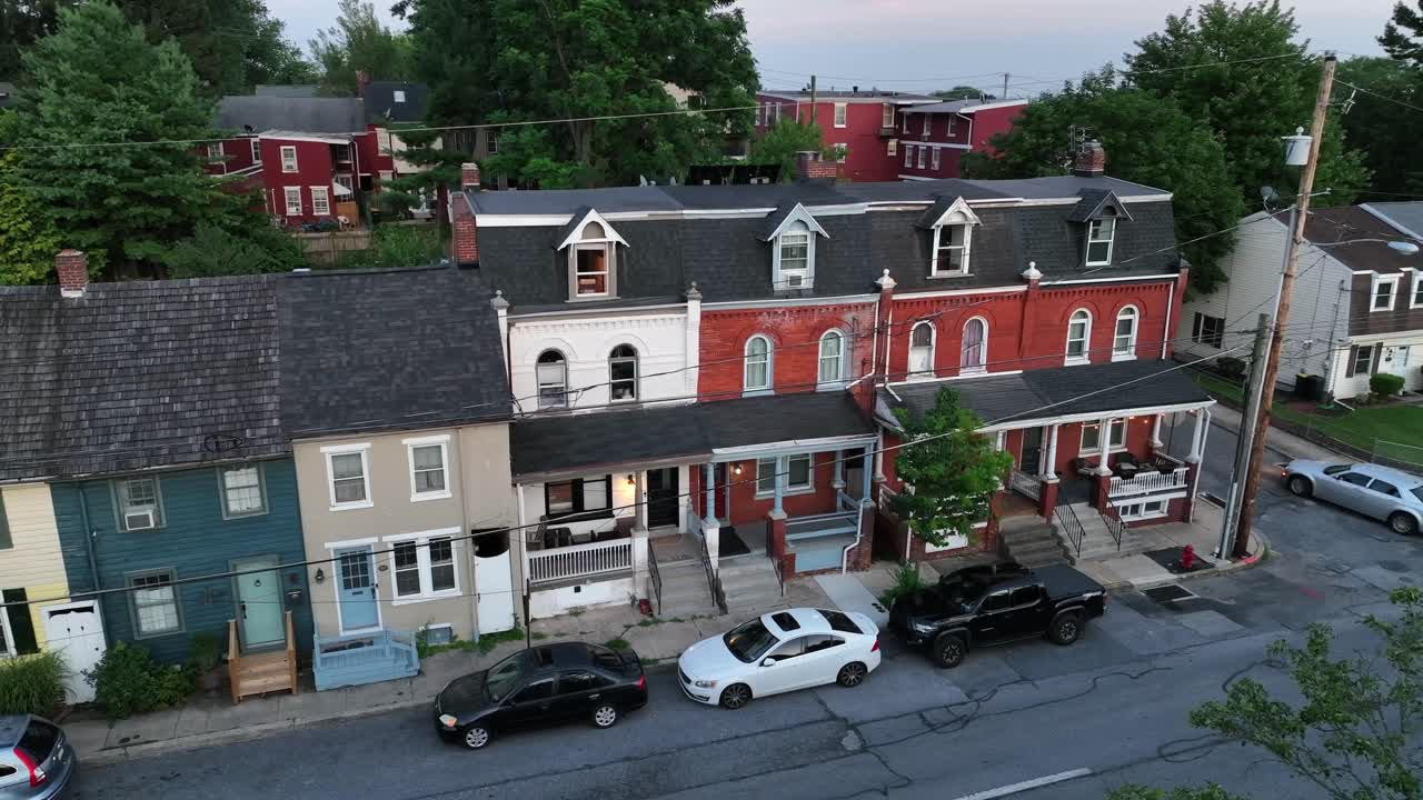 Aerial view of historic Pennsylvania rowhouses with colorful facades, brickwork and Mansard roofs, lined along quiet residential street with parked cars at dusk. Lateral shot. Porch and stairs