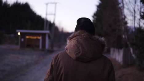 Footage-from-the-back-of-a-man-walking-down-the-countryside-road-on-cold-autumn-afternoon.-Wearing-black-hat-and-winter-coat-with-the-natural-fur-hood.-Evening-dusk.-Small-hut-on-the-background