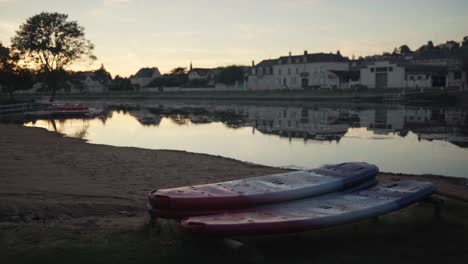 paddle boards sit idle next to a peaceful river and small village as evening closes in