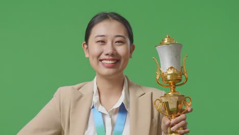 close up of asian business woman in a suit with a gold medal and trophy smiling to camera with one arm akimbo posing as the first winner on green screen background in the studio