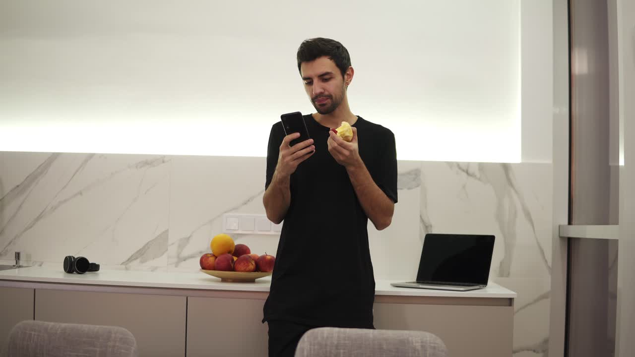 hombre guapo navegando en el teléfono móvil en casa cocina brillante. hombre joven guapo navegando en el teléfono inteligente sonriendo feliz. un hombre come una manzana