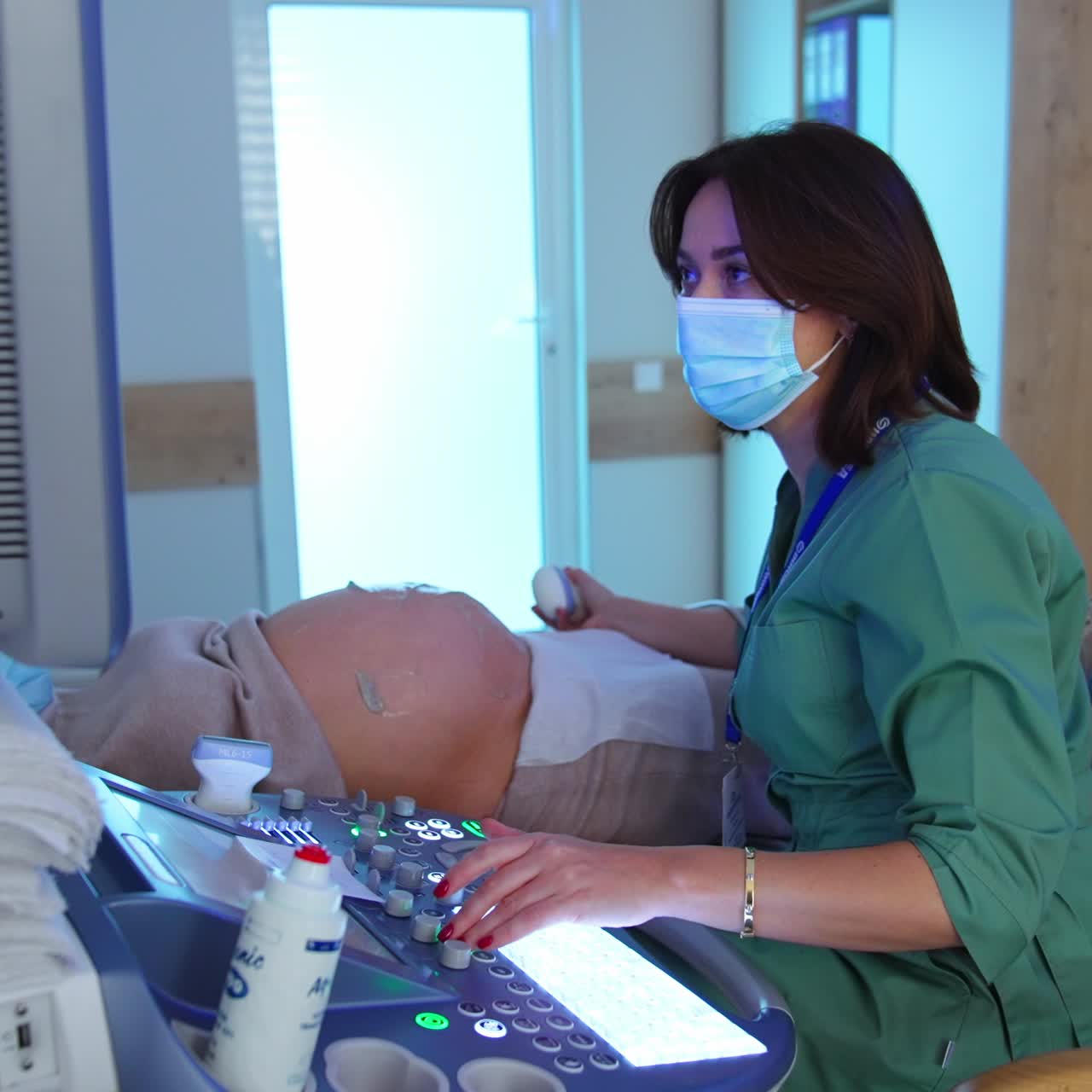 Female obstetrician sitting at the ultrasound equipment near pregnant lady. Doctor holding device, pressing buttons and commenting