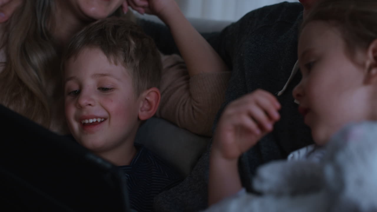 태블릿 컴퓨터를 사용하는 가족 어머니와 아버지는 아이들이 터치 스크린 장치에서 게임을 하는 것을 보고 재미를 배우고 잠자리에 들기 전에 양질의 시간을 즐기고 있습니다. 4k