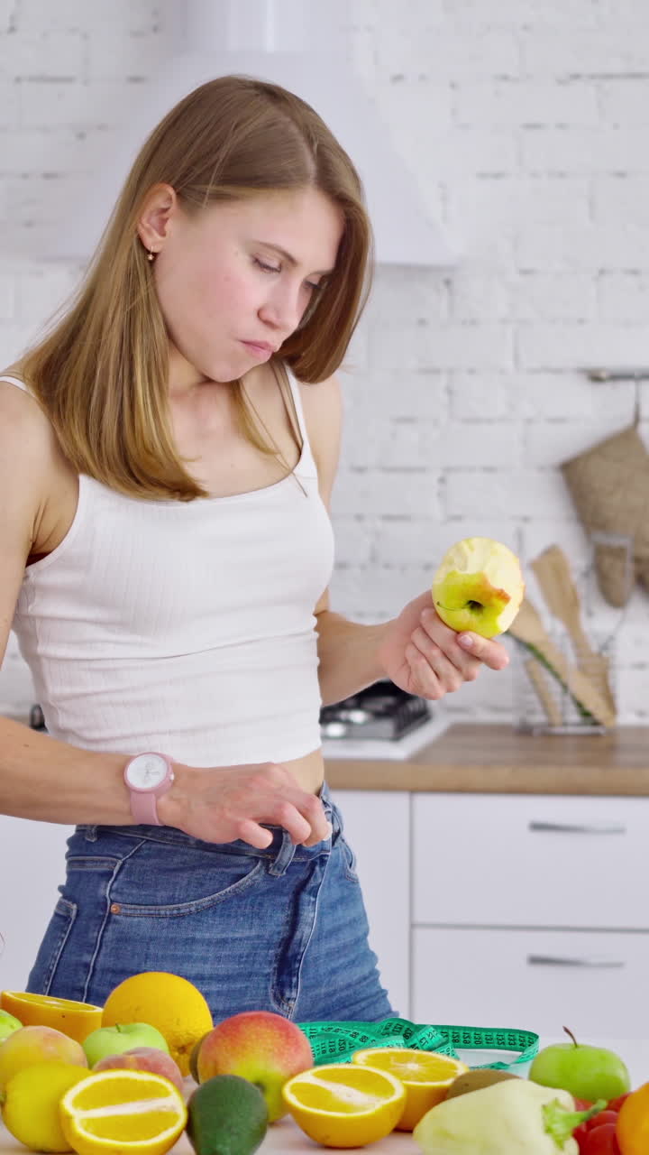 Losing weight concept. Young woman eating fruit in the kitchen. Beautiful model in jeans standing at the table full of organic vitamins. Vertical video
