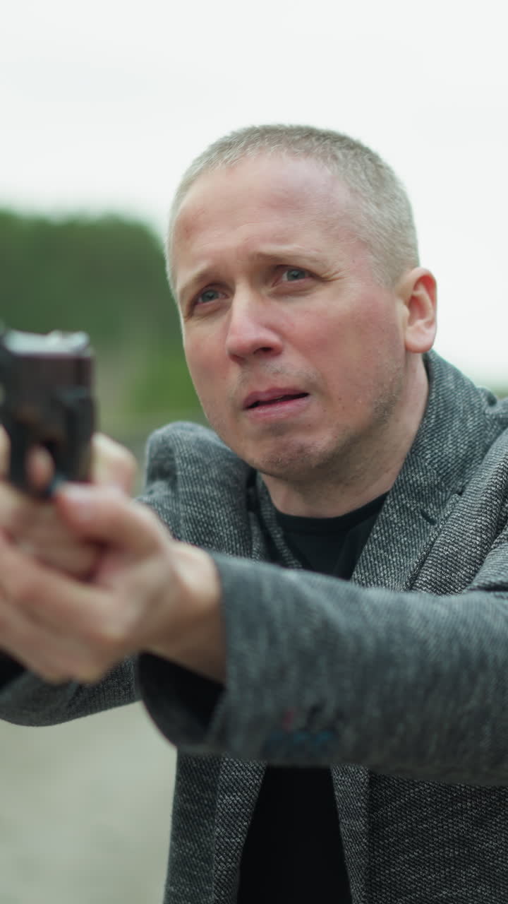 회색 재을 입은 남자가 확고한 표정으로 권총을 겨냥하는 근접한 모습, 배경은 흐릿한 철도 트랙과 숲을 보여줍니다.