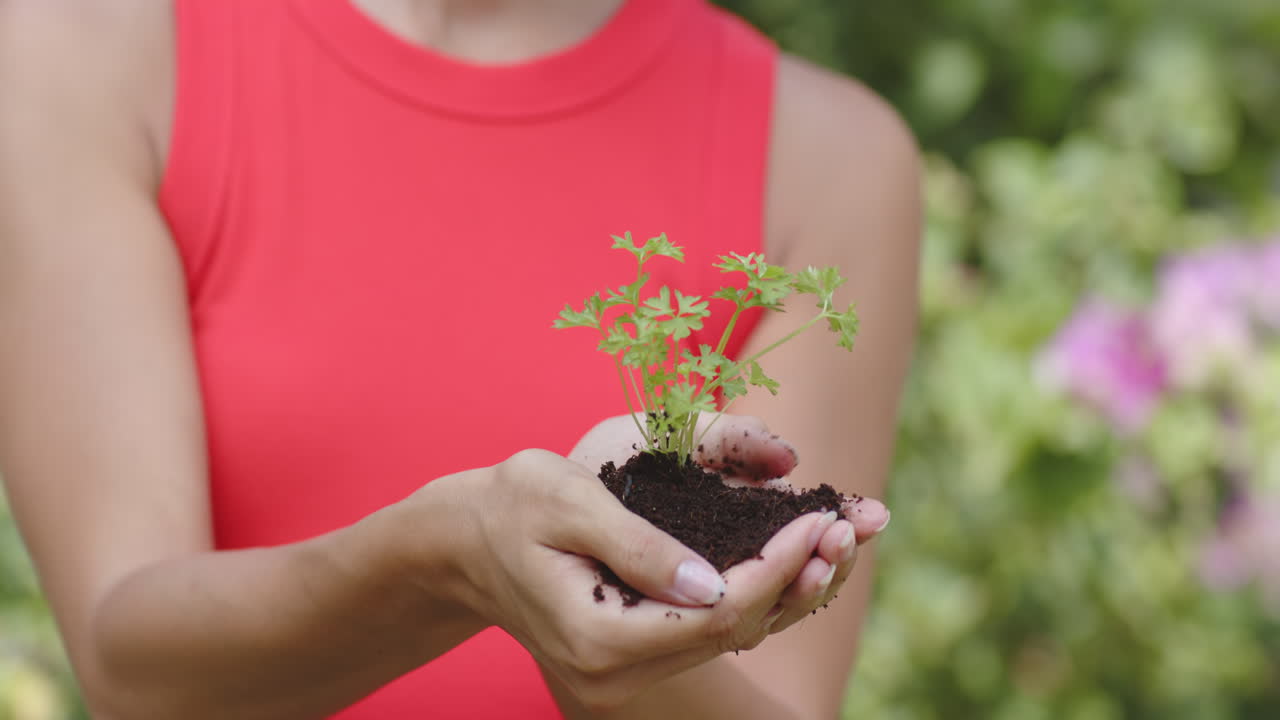 Smiling woman in red top holding small plant in garden, enjoying nature