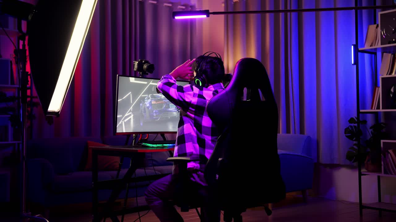 vista trasera de un streamer asiático que se estira mientras juega a un juego de carreras de coches a través de la red en una computadora personal. videojuego de transmisión en vivo, escritorio iluminado por luz de tira led rgb