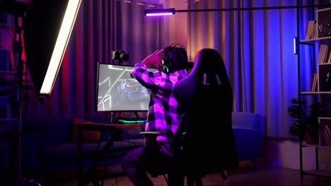 vista trasera de un streamer asiático que se estira mientras juega a un juego de carreras de coches a través de la red en una computadora personal. videojuego de transmisión en vivo, escritorio iluminado por luz de tira led rgb