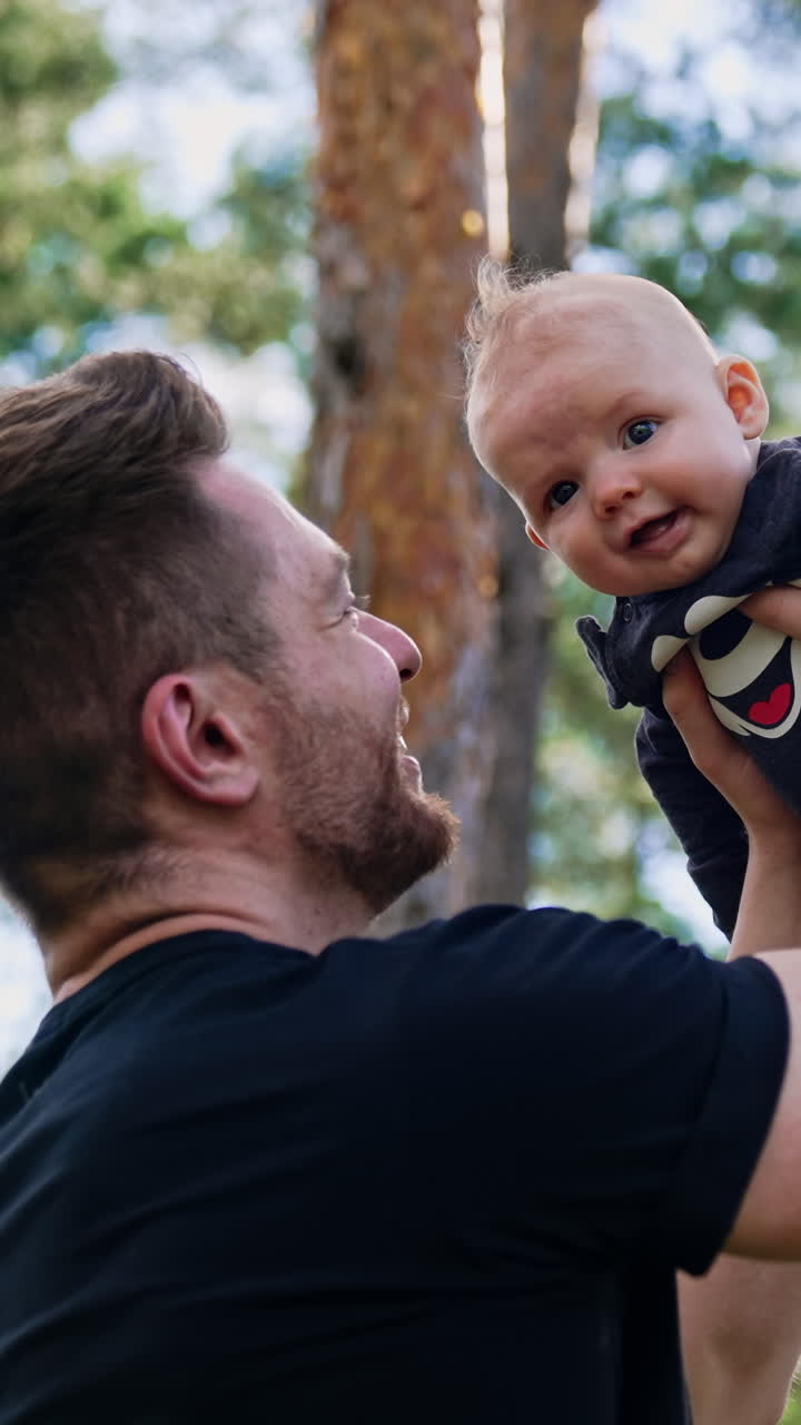 Bearded Caucasian man holding a cute newborn boy over the head. Happy dad looks with adoration on his baby. Low angle view. Pine trees at backdrop. Vertical video